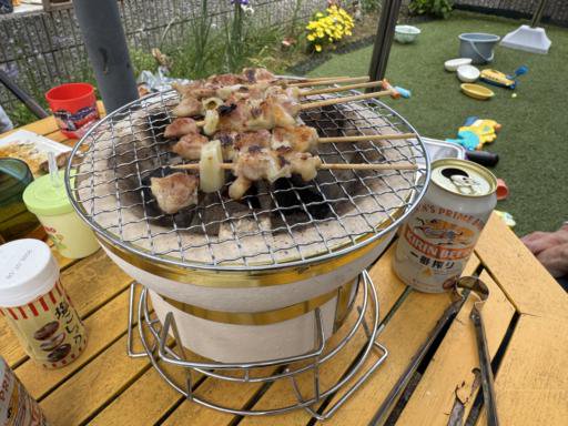 A small portable grill on a wooden table with skewered meat cooking. Nearby are a can of Kirin beer, metal tongs, and condiments in containers. Playground toys and balls are visible on the grass in the background, with yellow flowers along a brick wall.