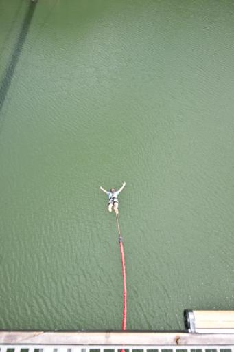 A person is captured in mid-air, arms extended, during a bungee jump over a large body of green water. The bungee cord, red in color, stretches from their harness to the platform at the bottom of the image. The waters surface appears slightly rippled.