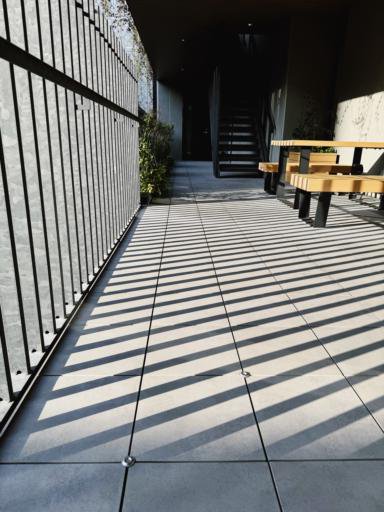 A corridor with a tiled floor casts diagonal shadows from a slatted fence on the left. A wooden bench with a black metal frame is on the right. In the background, there is a staircase leading upwards. Sunlight illuminates the scene, and some plants are visible along the left wall.