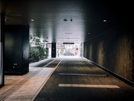 A covered walkway with a dark ceiling and walls leads to an urban street. The path has textured flooring, with tactile paving lines. To the left, there