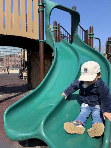 A child wearing a light-colored cap, dark jacket, and jeans slides down a curved, green double slide at a playground. A bright blue sky and a building are visible in the distance.